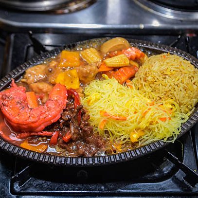 A close-up of an assorted meal featuring fried rice, stir-fried noodles, shrimp, beef sauce, and mixed vegetables served on a black plate. A fusion of Nigerian and Asian-inspired flavors presented on a stovetop